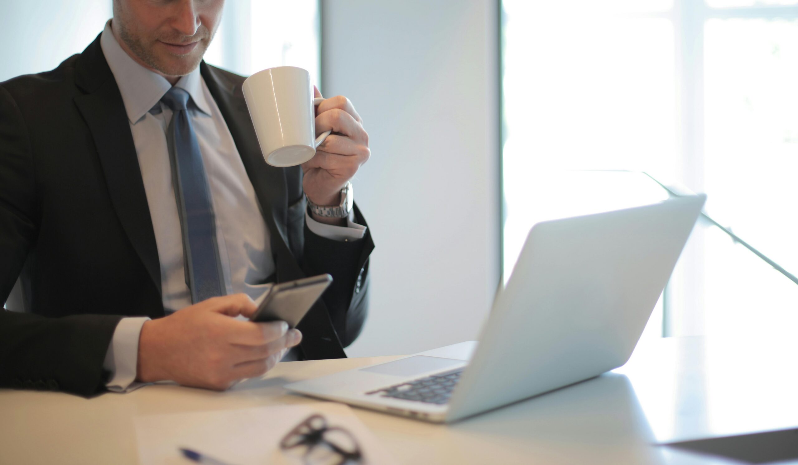 Man in a suit drinking coffee and looking at his phone in front of a laptop